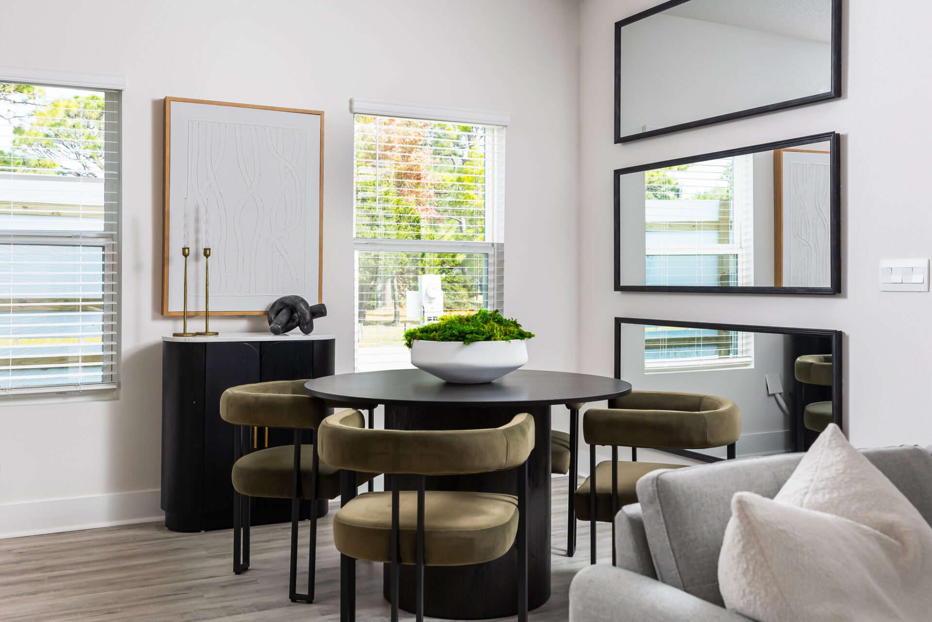 A stylish dining area with a round black table and four green velvet chairs. A white ceramic planter with green moss serves as a centerpiece. Large mirrors and modern art pieces decorate the wall.