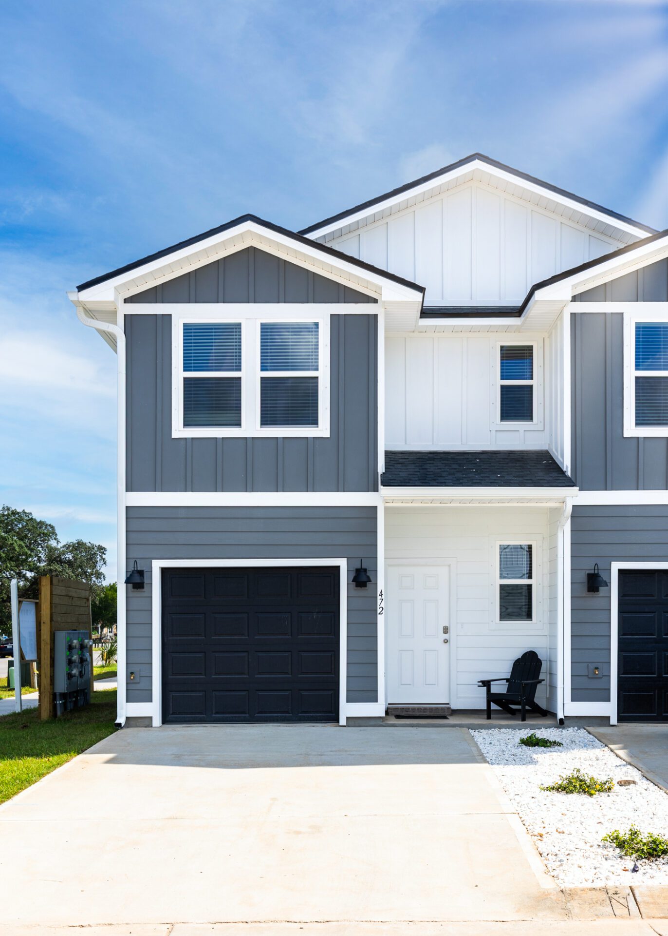 The front view of a two-story modern home with gray and white siding, a white front door, and black garage doors. There are two black chairs in front of the entrance.