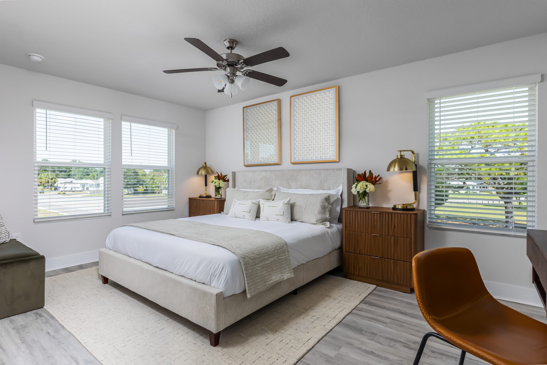 A wide-angle view of the master bedroom featuring a beige upholstered bed, wooden nightstands with gold lamps, and large windows with white blinds. The room has a neutral color palette with subtle geometric decor.